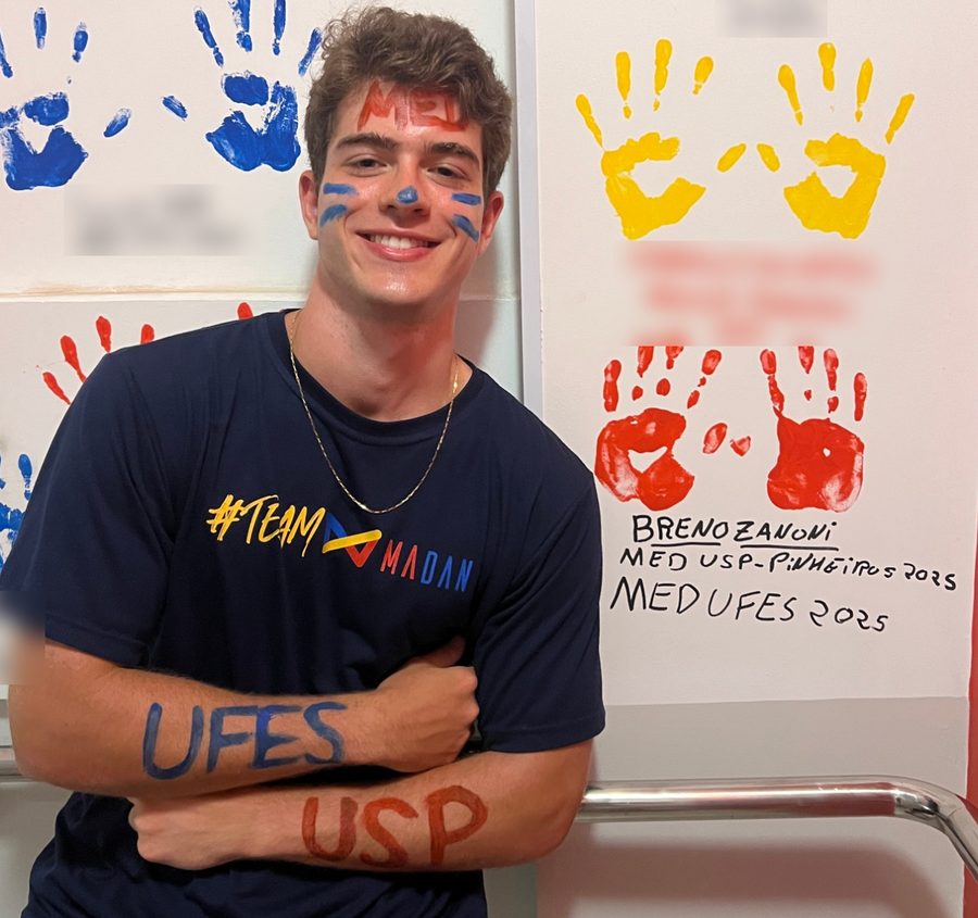 Breno Zanoni no mural de aprovados, com USP e UFES pintados nos braços e as mãos assinadas: MED USP-Pinheiros 2025 e MED UFES 2025